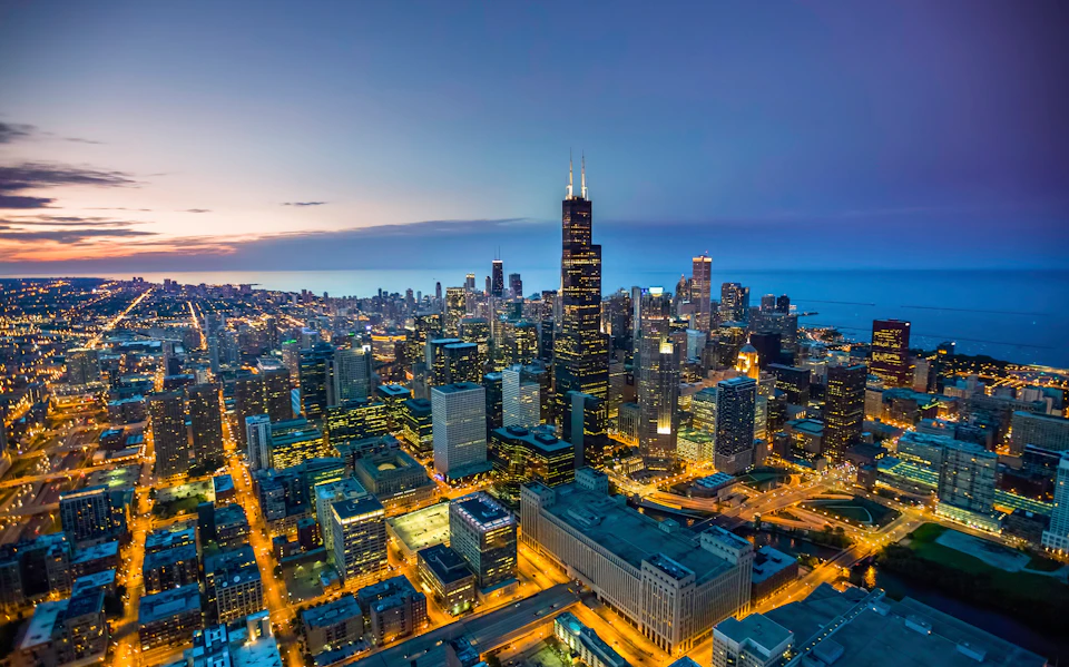 Skyline view of a city at dusk with illuminated skyscrapers and streets, and a large body of water in the background.
