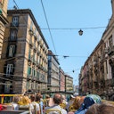 People seated on the upper deck of an open-top bus, traveling through narrow city streets lined with tall, colorful buildings.