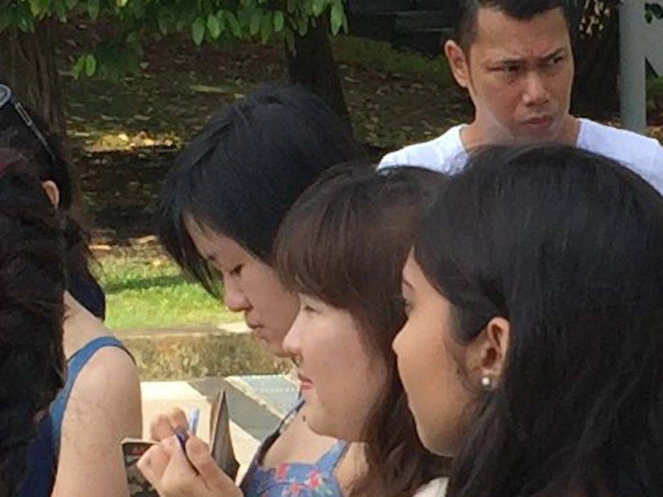 People standing outdoors in a park, some chatting and others looking around. Greenery and a paved path are visible.