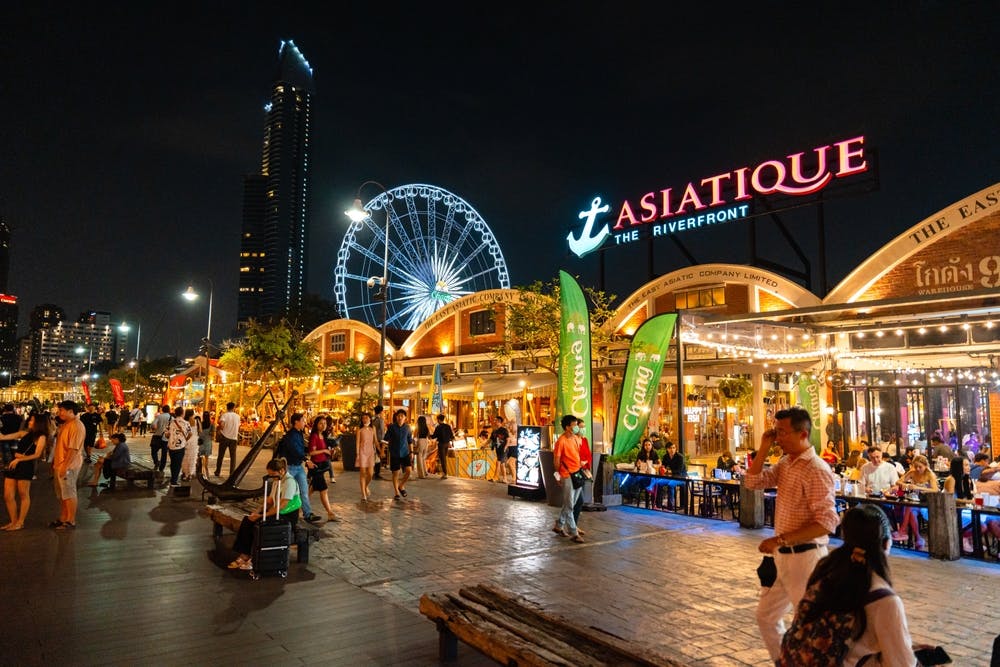 Night scene at Asiatique The Riverfront, bustling with people, bright lights, shops, eateries, and a Ferris wheel in the background.