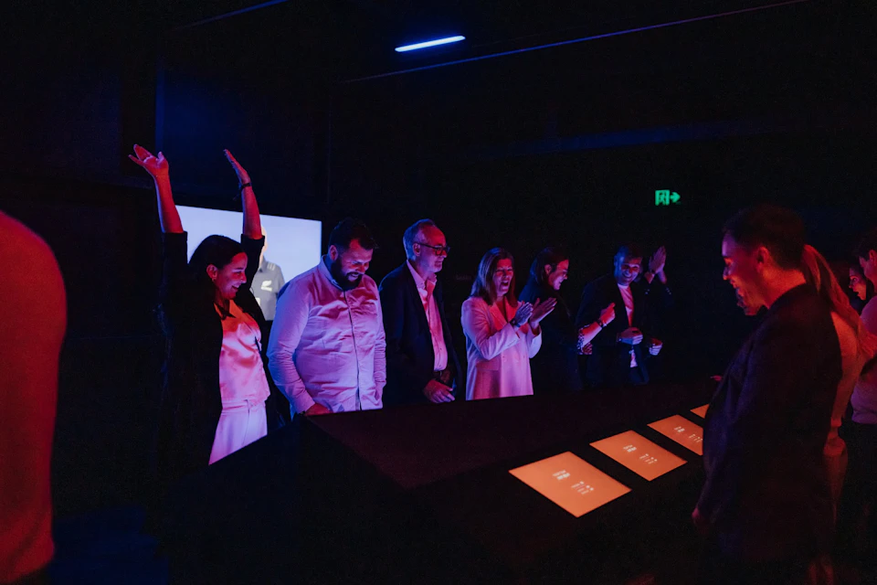 A group of people in a dark room interacting with illuminated touchscreens, under colored lighting.