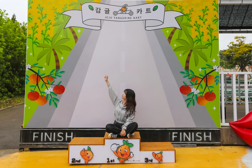 A person sits on a podium decorated with cartoon oranges in front of a backdrop featuring palm trees and a road.