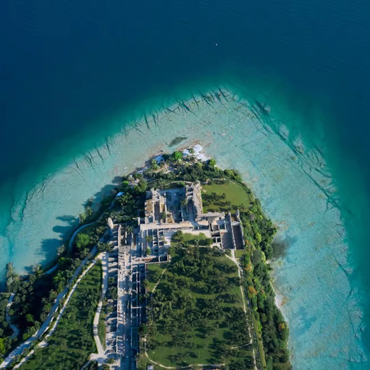 Aerial view of an island with ruins, greenery, and surrounding turquoise and deep blue water.