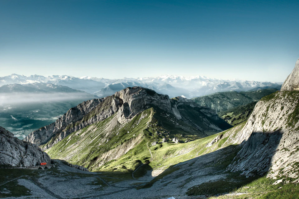 Vast mountainous landscape with rugged peaks, green valleys, and distant snow-capped mountains under a clear blue sky.
