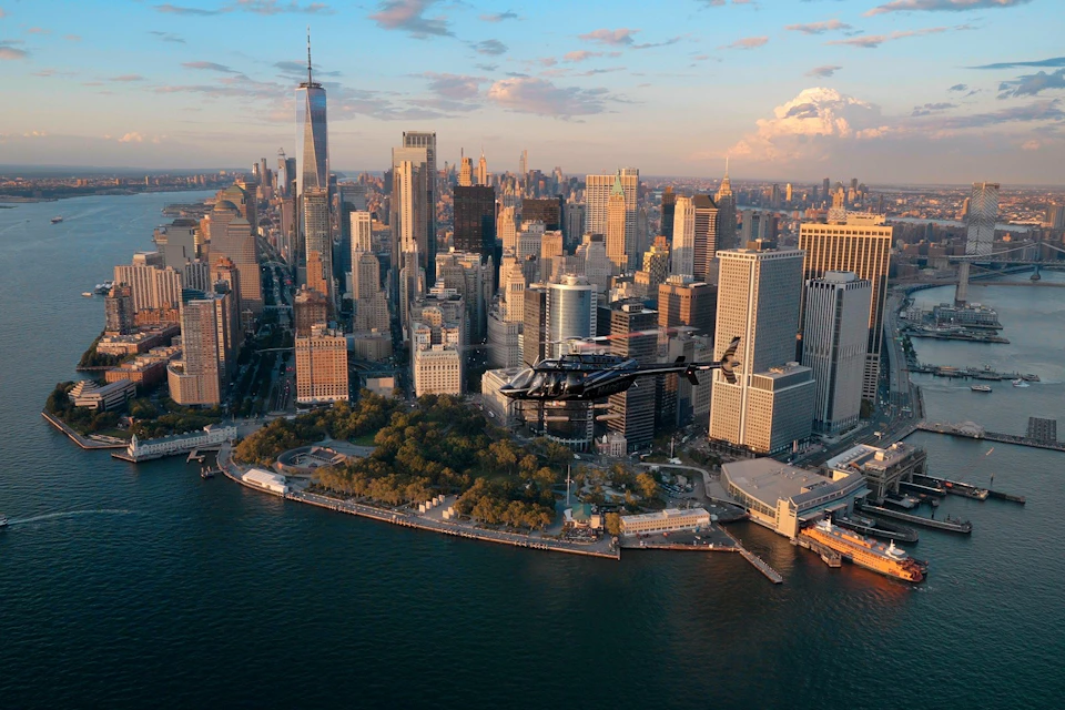 A helicopter flying over lower Manhattan with skyscrapers in the background and the waterfront in the foreground during sunset.