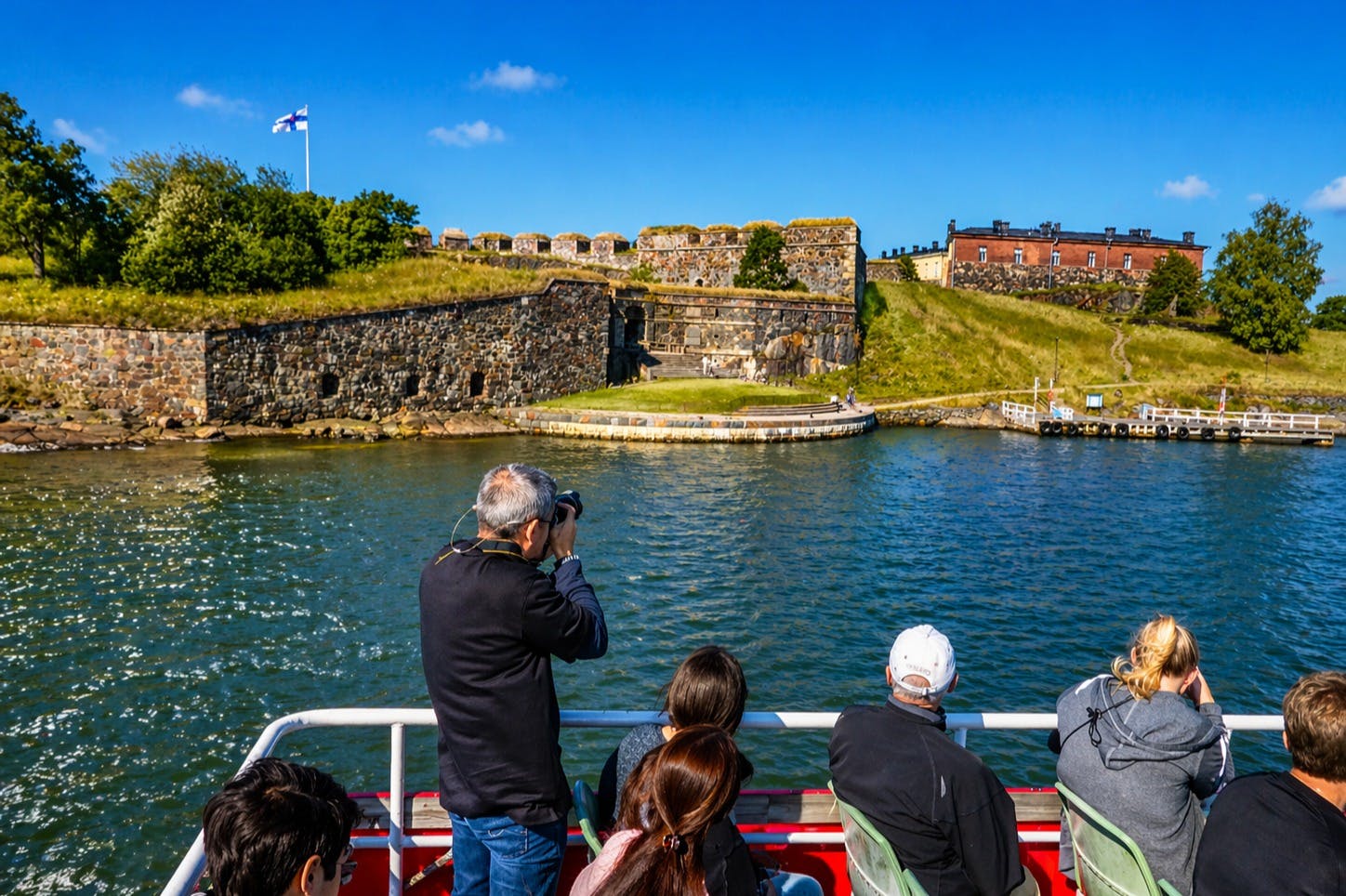 Gent en un ferri observant una fortalesa històrica i un paisatge herbós a través d'un canal d'aigua sota un cel blau clar.