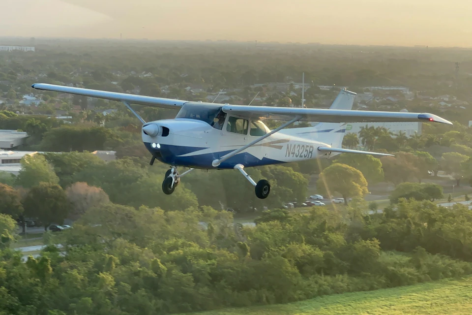 A small blue and white propeller airplane flying over a forested area at sunset, with the plane's registration number N4325R visible.