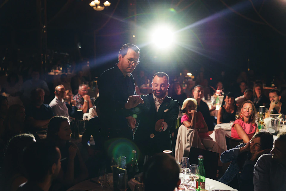 A man performs a magic trick for another man in a suit at a dimly lit event, while the seated audience watches intently.