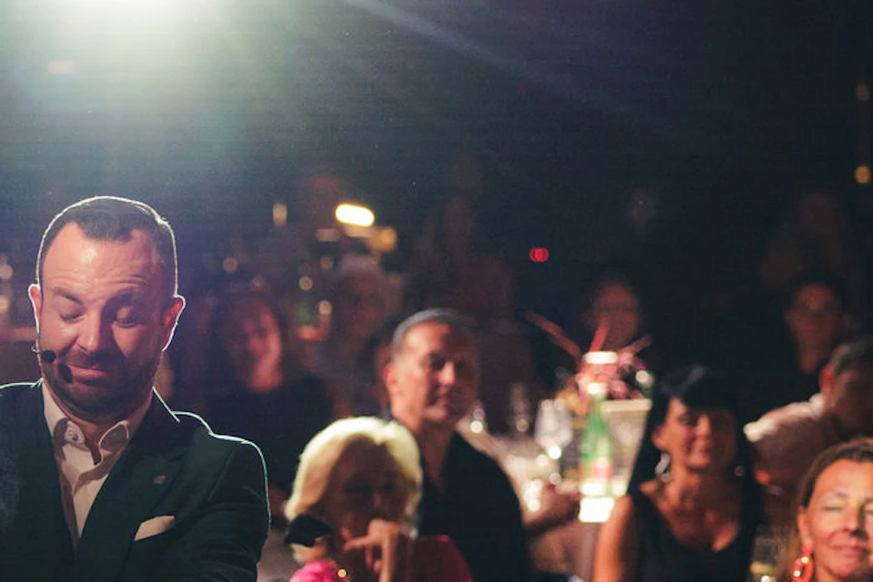 A man performs a magic trick for another man in a suit at a dimly lit event, while the seated audience watches intently.