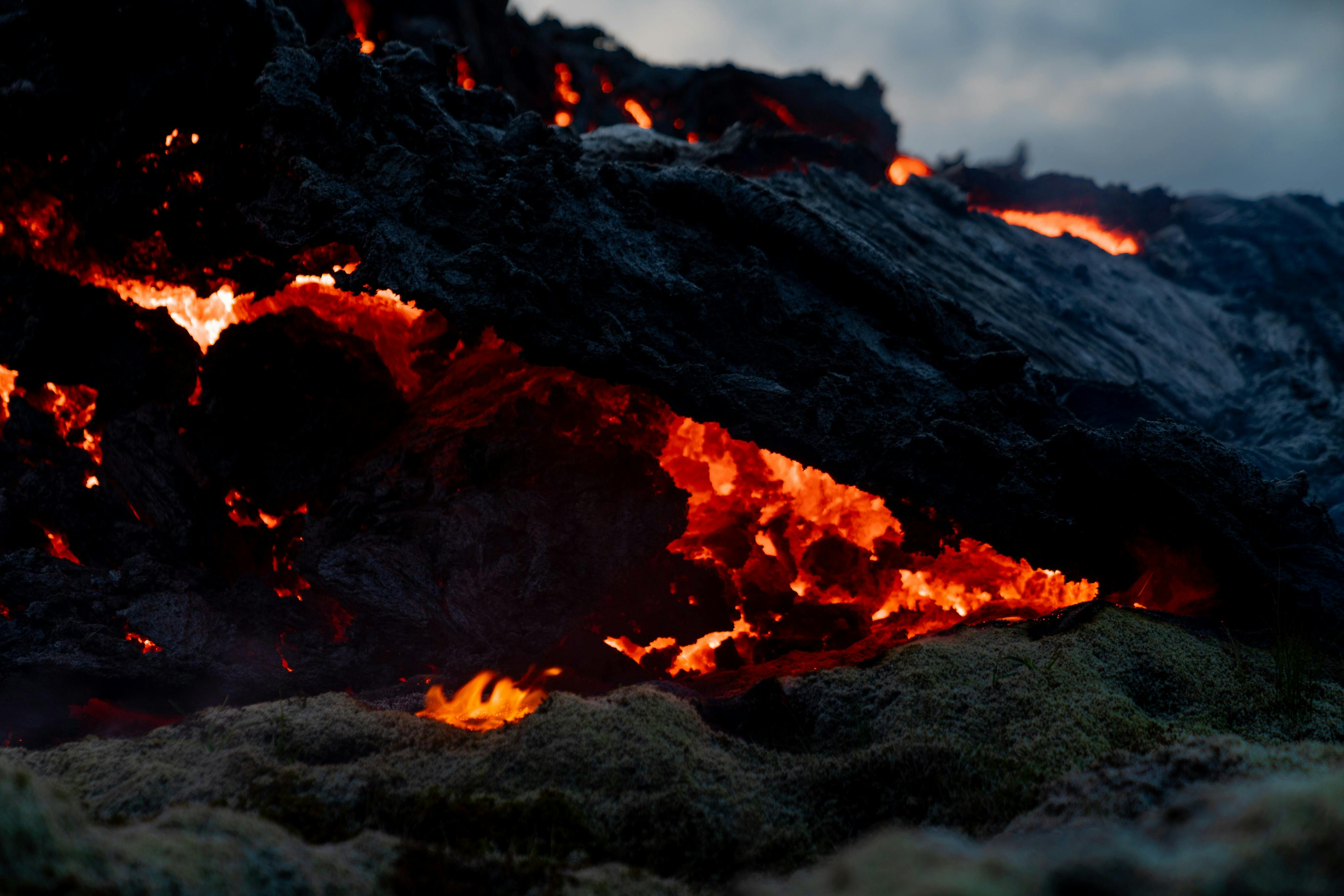 Расплавленная лава, светящаяся ярким оранжевым светом, течет под охлажденной темной вулканической породой под облачным небом.