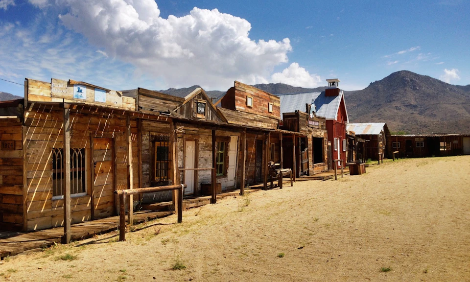 Old-West style town with wooden buildings, a dirt street, and a mountainous background under a partly cloudy sky.
