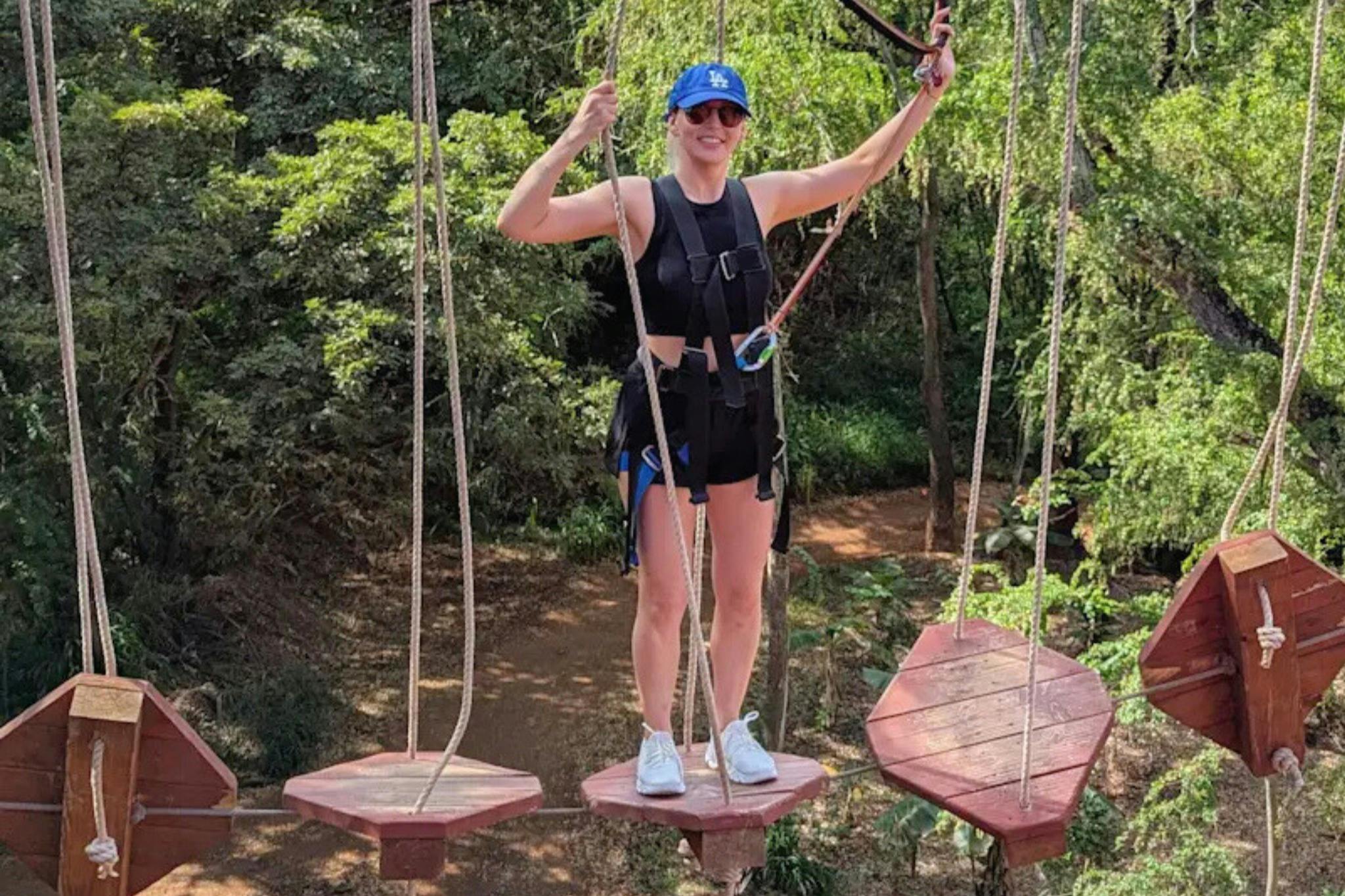 A person wearing a harness and helmet is on a rope bridge with wooden platforms, surrounded by trees and greenery.