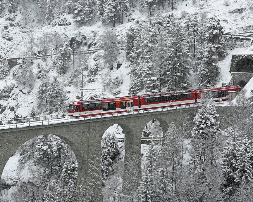 Ένα κόκκινο τρένο διασχίζει μια πέτρινη αψιδωτή γέφυρα μέσα από ένα χιονισμένο, δεντρόφυτο τοπίο.