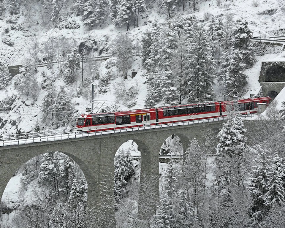 A red train crosses a stone arch bridge through a snowy, tree-covered landscape.