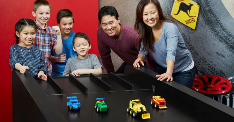 Children and adults excitedly watch a race of colorful LEGO car models on a black track.