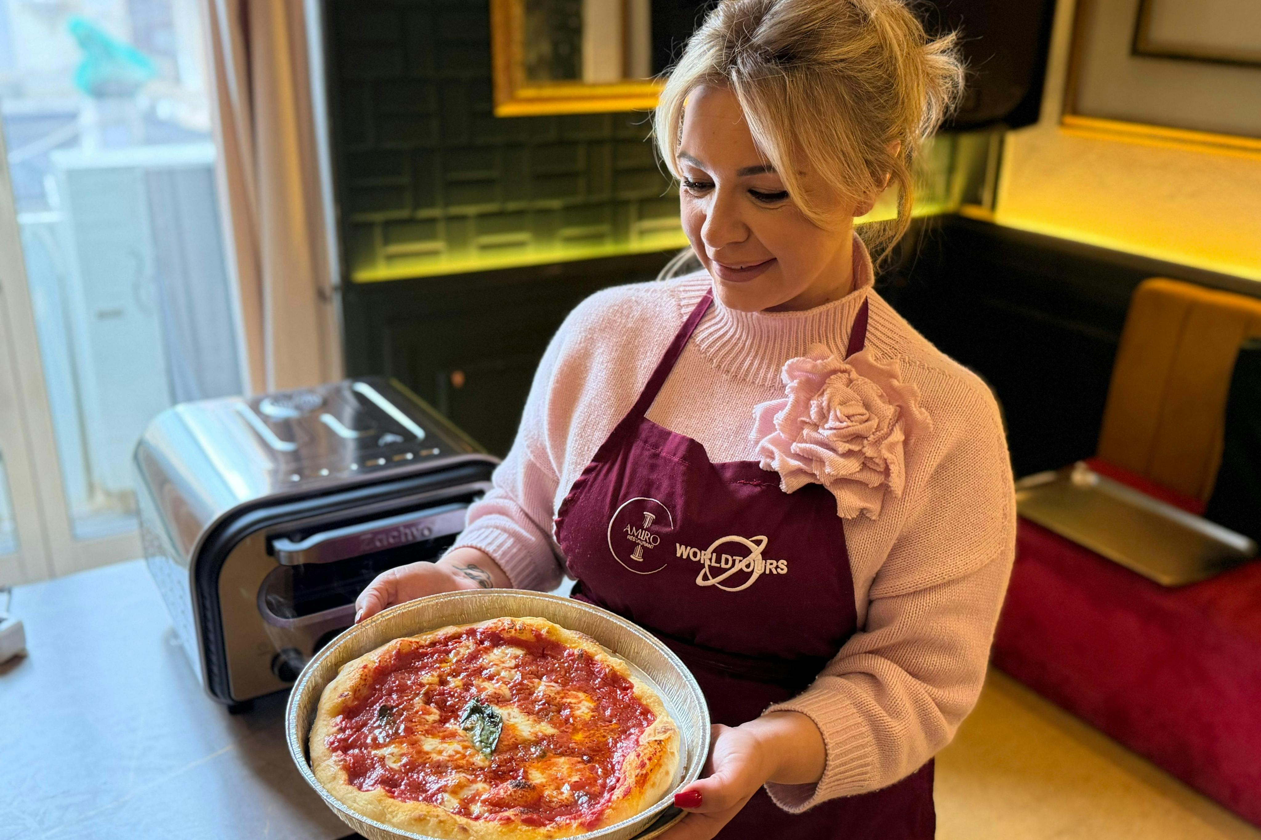 A woman wearing a pink sweater and maroon apron holds a plate with a pizza in a cozy kitchen setting.