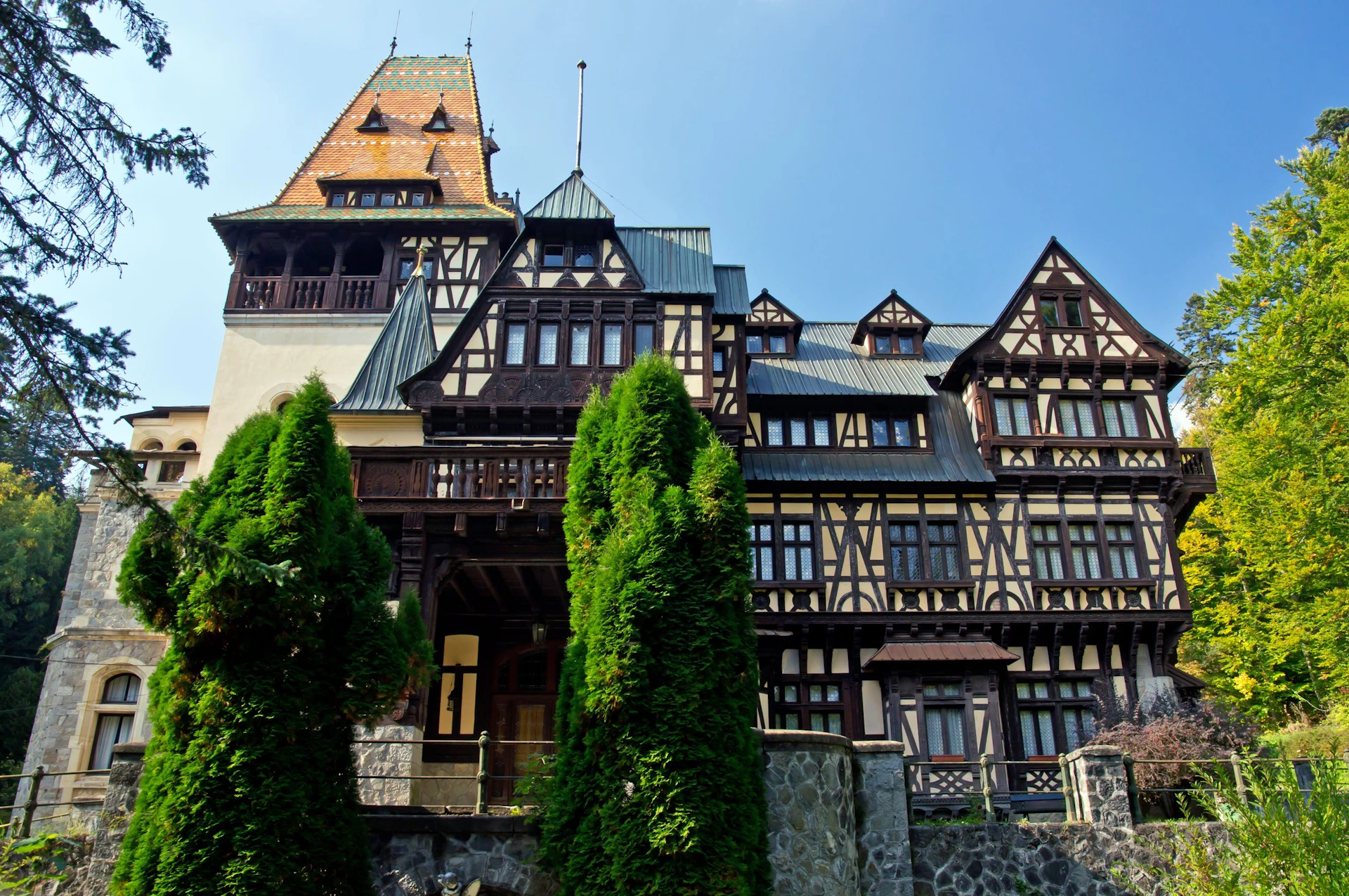 A multi-story building with a steep roof, numerous windows, and timber-frame design, flanked by tall conifer trees.
