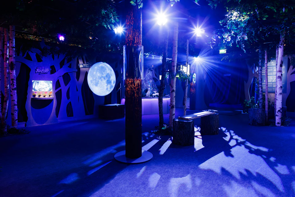 A dimly lit, indoor forest-themed space with artificial trees, a large moon backdrop, blue lighting, and tree stump seating.