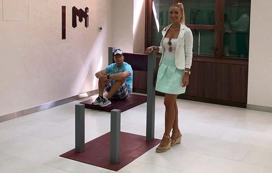 Vedle optické iluzorní struktury stojí usměvavá žena a v pozadí na plošině sedí zdánlivě muž.