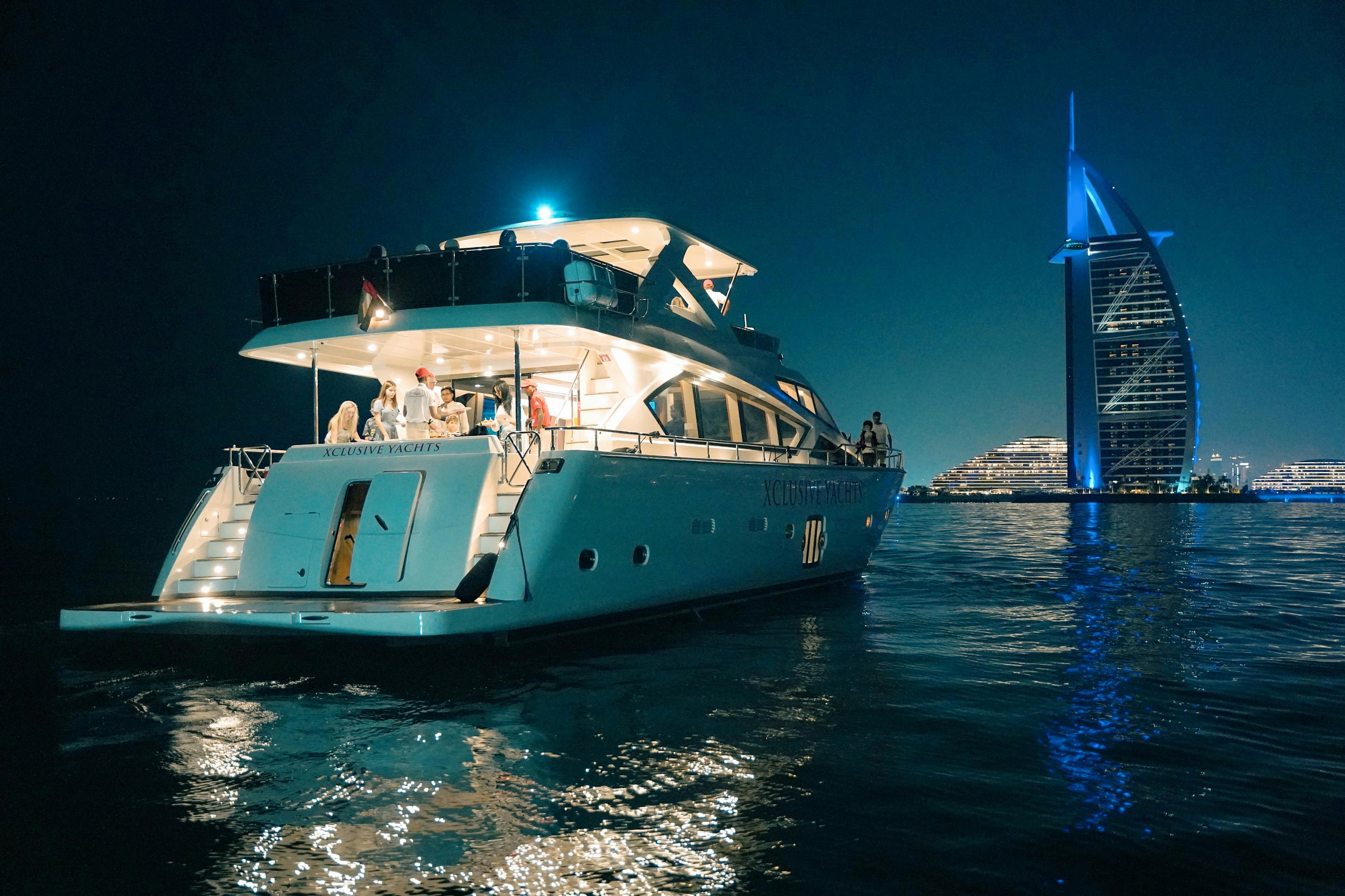 Eine nächtlich beleuchtete Yacht mit Menschen auf ihrem Deck in der Nähe des Burj Al Arab im Hintergrund.