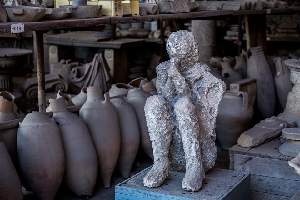 Ancient plaster cast of a crouching figure amidst old pottery and artifacts on shelves in a historical setting.