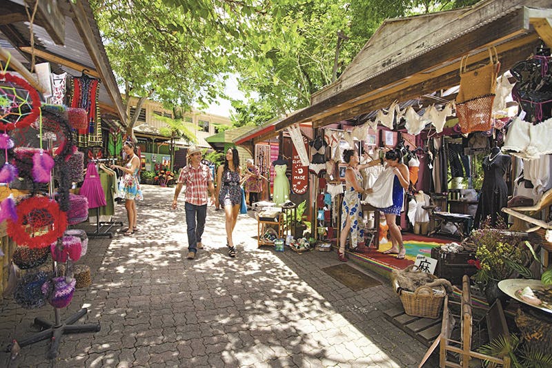 People shopping and walking along a tree-shaded outdoor market with various clothing stalls and vibrant merchandise displays.