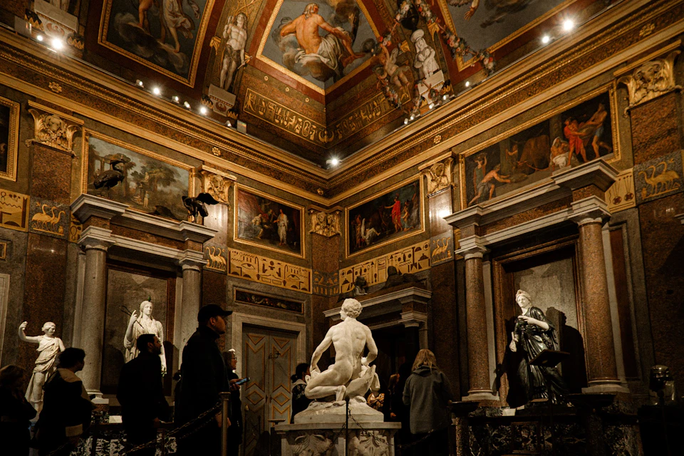 Visitors observe classical sculptures in an ornate, frescoed museum room with detailed ceiling paintings and columns.