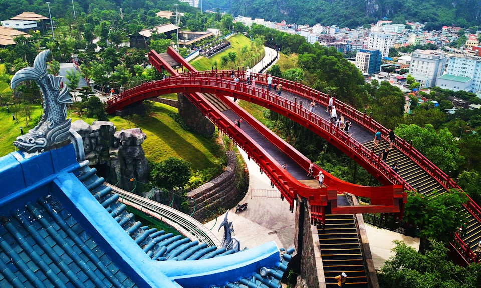 A scenic red pedestrian bridge curves over a lush landscape with visitors walking and a city visible in the background.