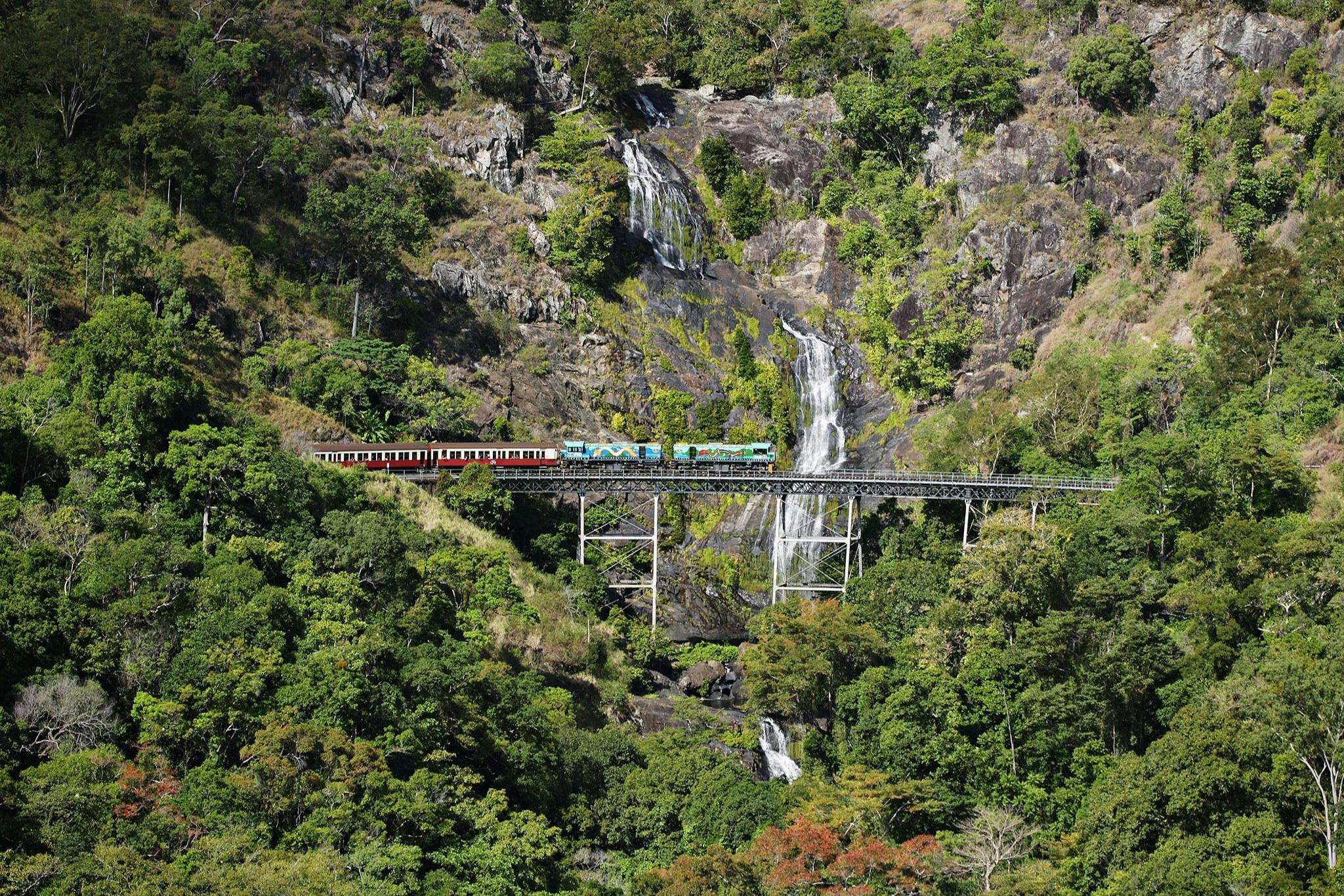A train travels on a bridge surrounded by lush green forest and a cascading waterfall in the background.