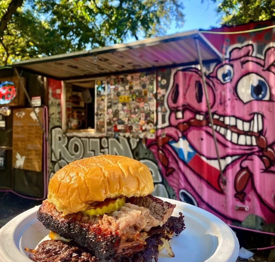 A brisket sandwich on a plate in front of a graffiti-covered food truck with a pig mural.