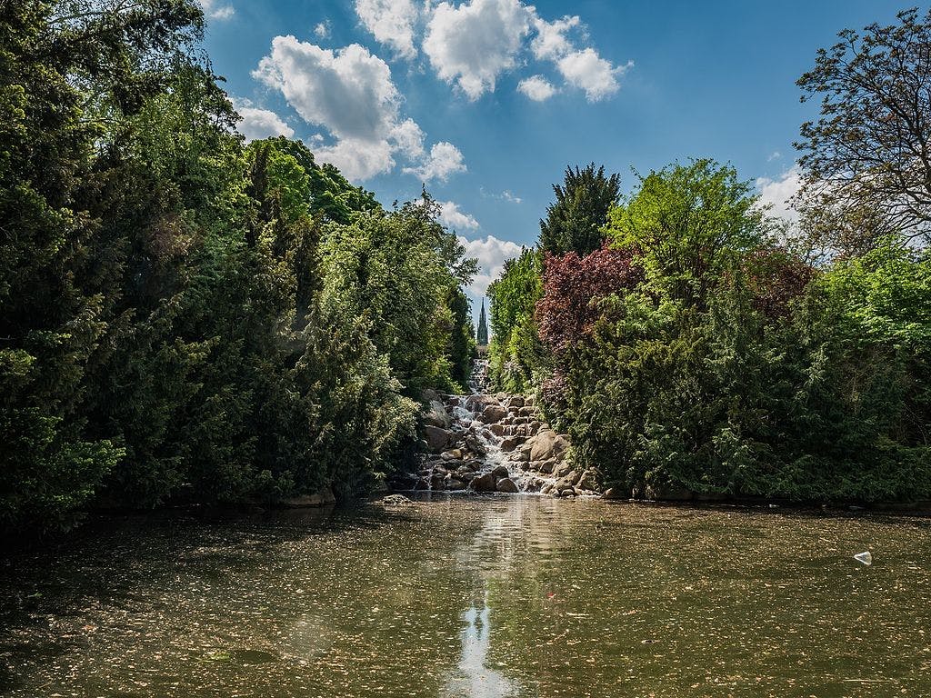 A serene scene of a small waterfall surrounded by lush greenery, cascading into a calm pond under a blue sky dotted with clouds.