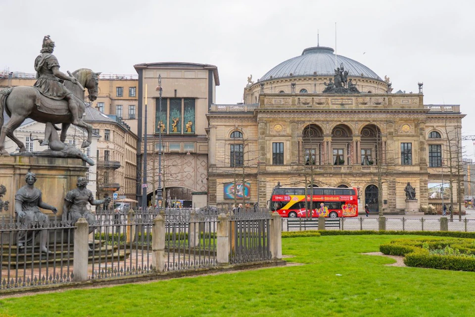A city scene with historic buildings, a statue in a fenced area, and a red double-decker bus on the street.