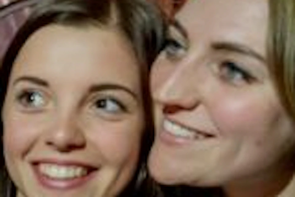Two young women take a selfie inside a lit bus or tram at night, smiling and posing with bright lights reflected on the windows.