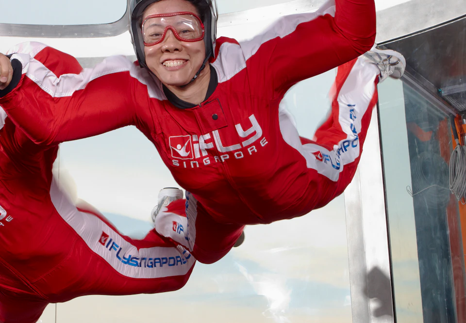 Two people in red jumpsuits are indoor skydiving inside a vertical wind tunnel. An operator stands nearby.