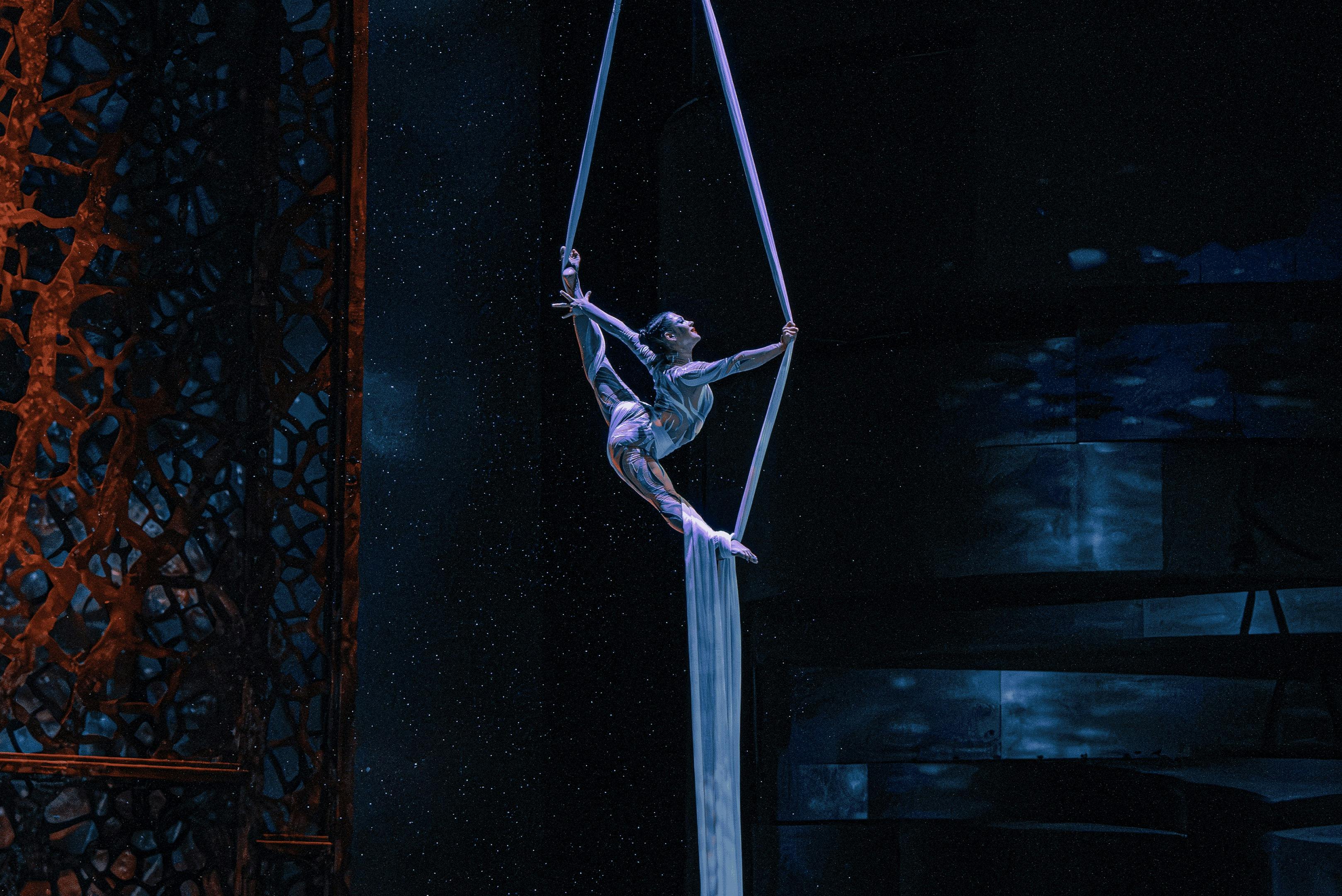 Een performer hangt in de lucht met behulp van vliegend zijde, omringd door een donker en sfeervol podium met subtiele verlichting.