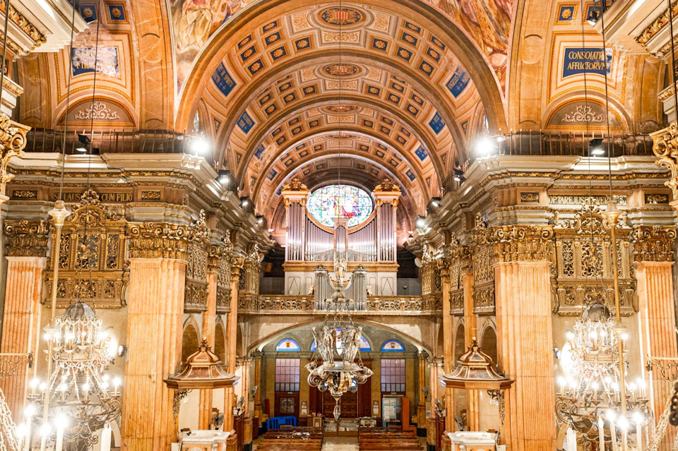 Interior of an ornate church featuring a decorated vaulted ceiling, chandeliers, and a large stained glass window.