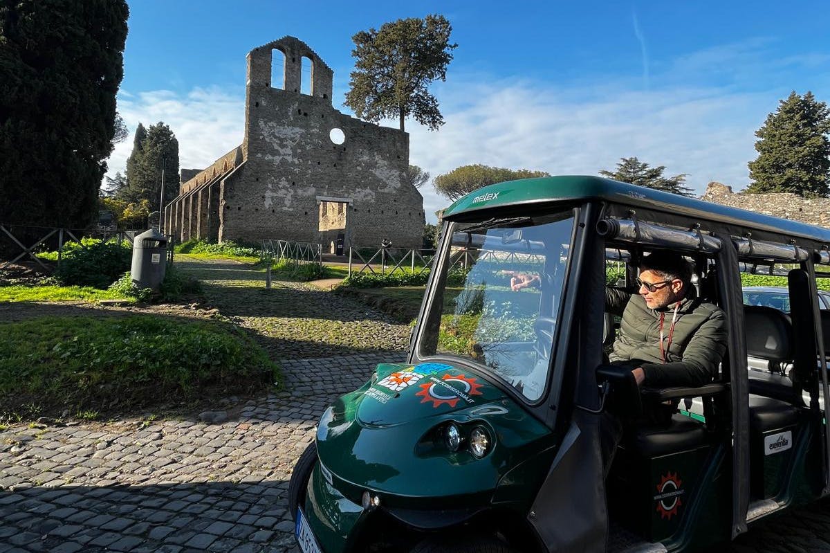 Un hombre está sentado en el interior de un carro turístico verde aparcado en un camino empedrado con ruinas antiguas y árboles al fondo.