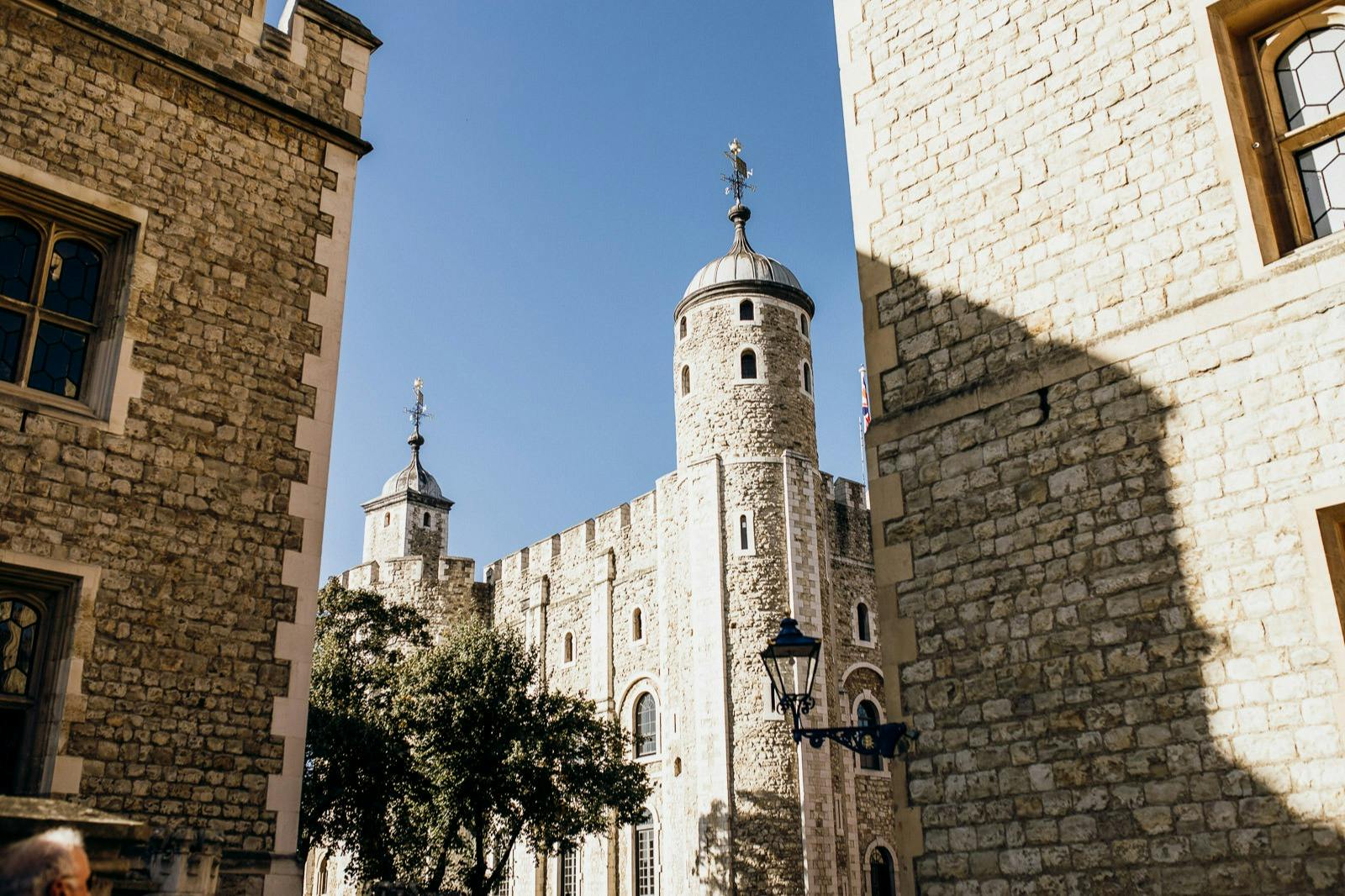Stone towers and walls of a historic building under a clear blue sky, flanked by trees and a vintage lamp post.
