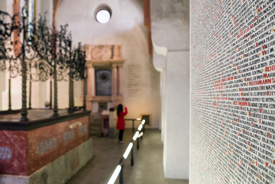 A person in a red coat takes a photo inside a historical building with a large wall covered in text and an elaborate wrought-iron structure.
