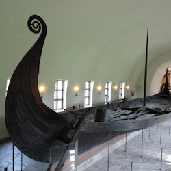 Ett gammalt vikingaskepp visas inomhus med en böjd bock, mast och bänkar. Besökare tittar på fartyget i bakgrunden.