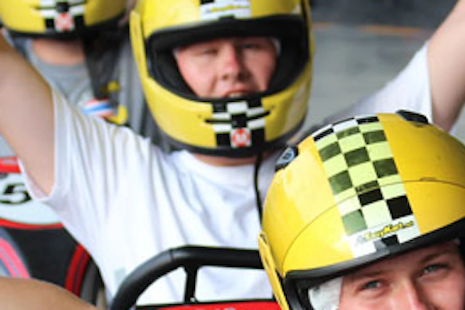 Three people wearing yellow helmets in go-karts, smiling and making peace signs. Attached numbers on karts are visible.