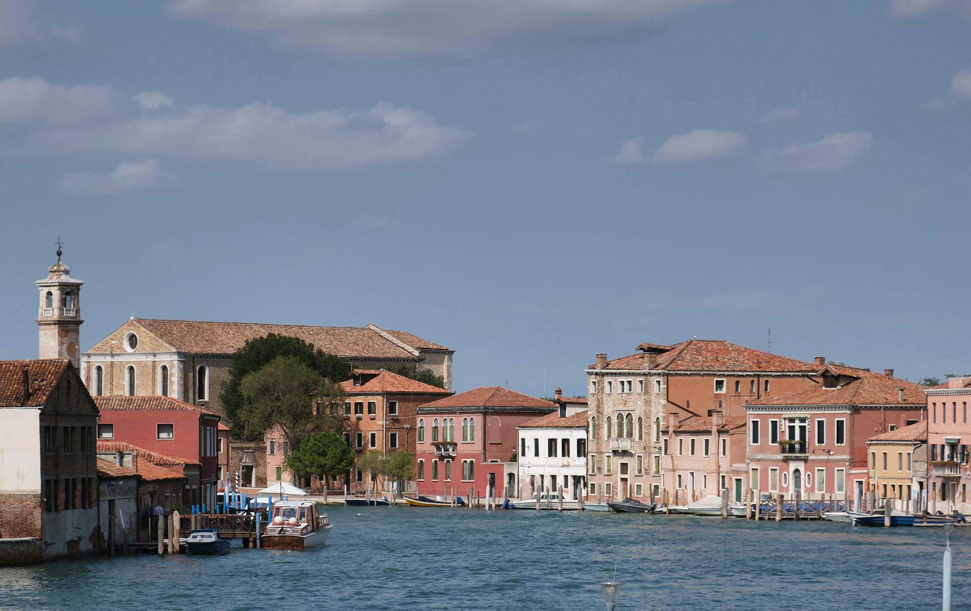 En kanal med färgglada byggnader, båtar dockade och en kyrka i bakgrunden under en blå himmel i Venedig.
