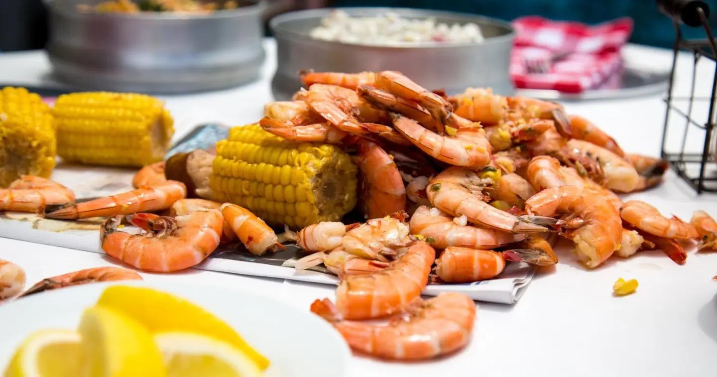 Une pile de crevettes cuites à côté d'un épi de maïs posé sur une table, avec des plats de nourriture visibles à l'arrière-plan.