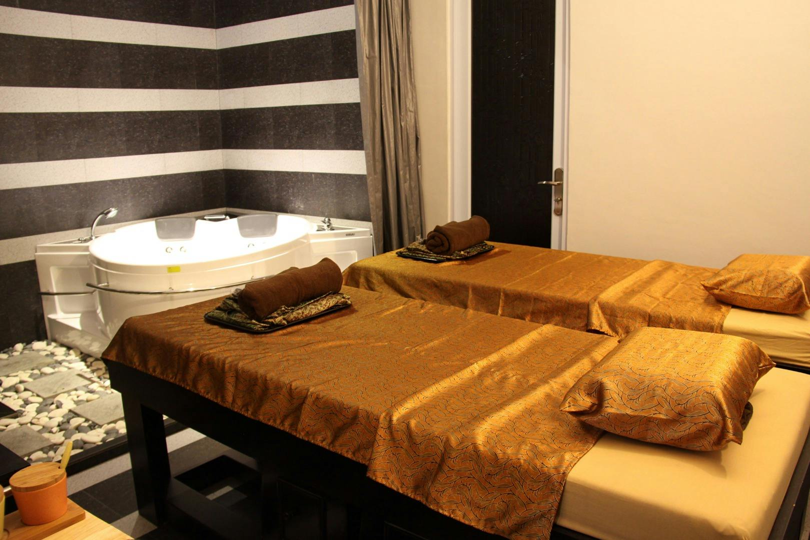 Pokój spa z dwoma stolikami do masażu pokrytymi złotą tkaniną, wanną jacuzzi i ozdobnymi kamieniami na podłodze.
