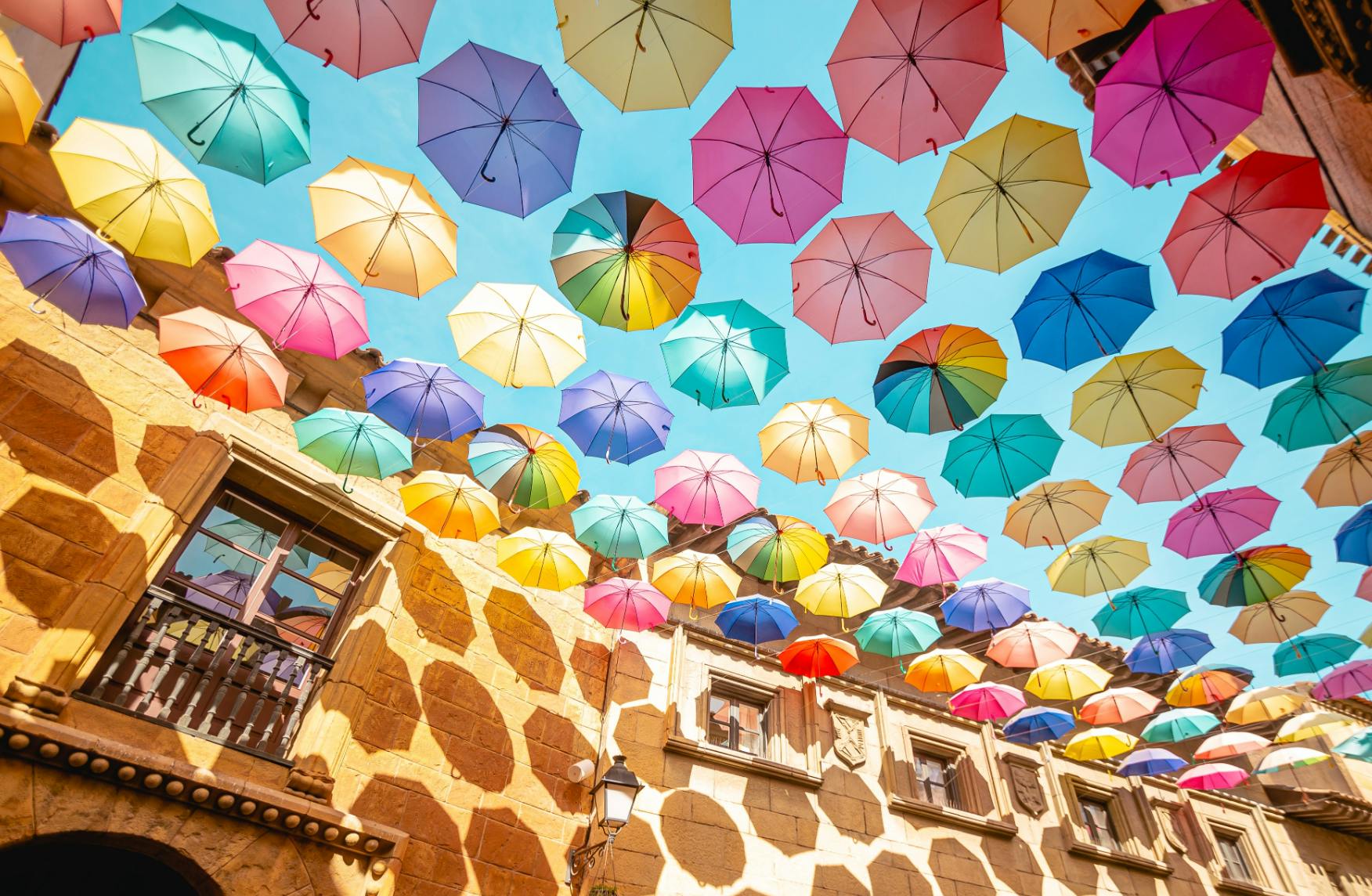 Kolorowe parasole zawieszone nad dziedzińcem z kamiennymi zabudowaniami i błękitnym niebem.