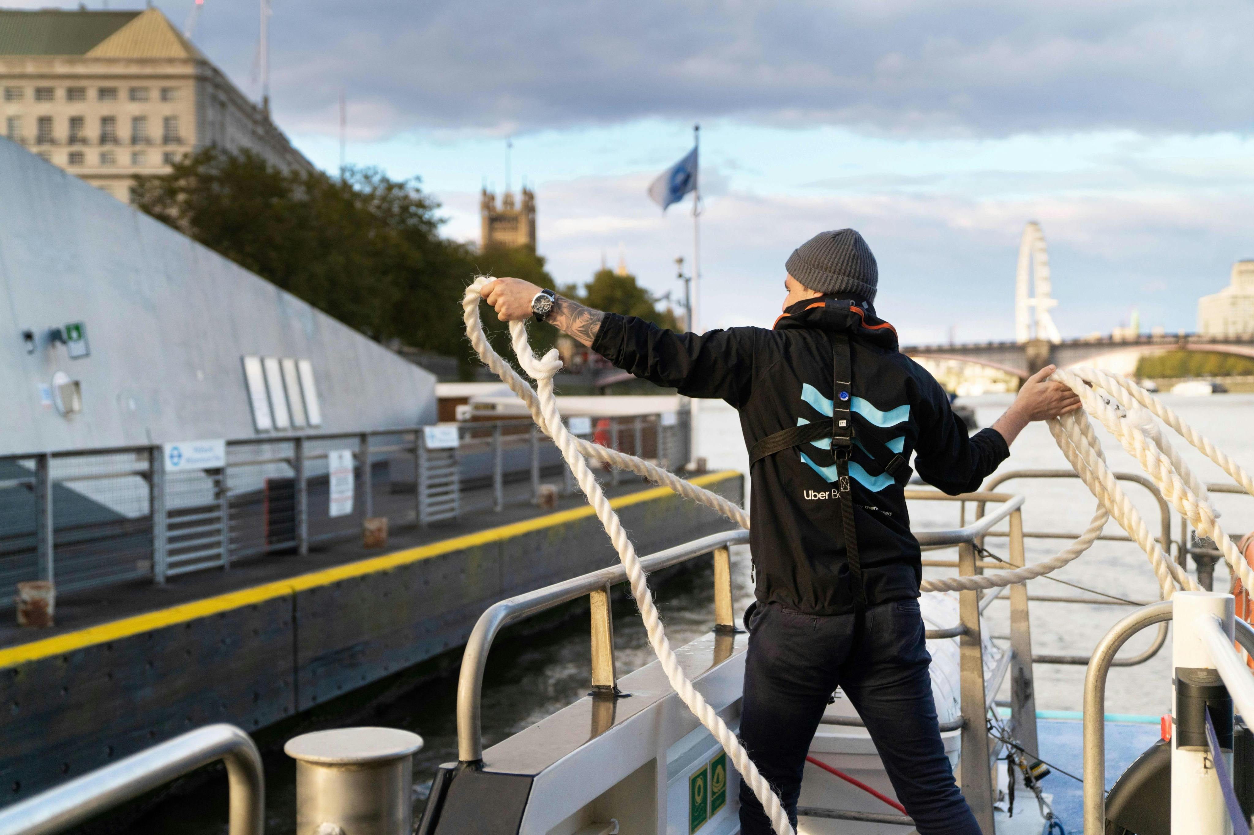A person on a boat throwing a rope towards a dock, with a cityscape and a ferris wheel visible in the background.