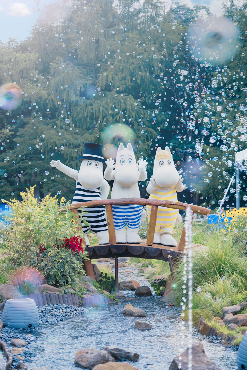 Three cartoonish characters in striped clothing stand on a wooden bridge in a garden with bubbles floating around.