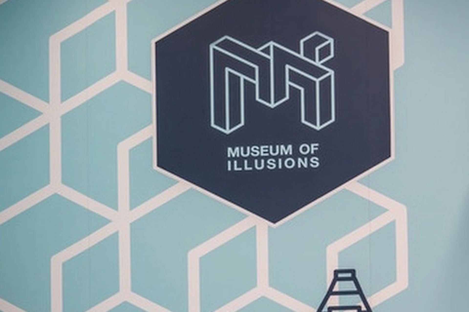 Two people in a room with a geometric pattern and "Museum of Illusions" sign; one appears larger due to perspective.
