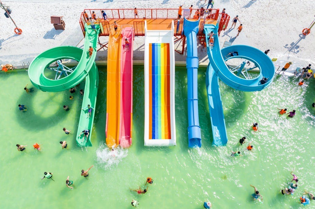 People enjoying various colorful water slides and swimming in a pool, viewed from above on a sunny day.