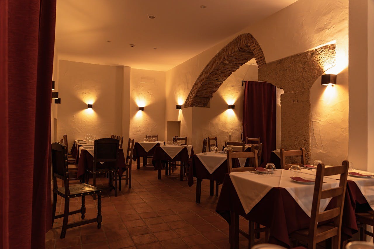 Mírně osvětlená restaurace s klenutými kamennými dveřmi, bílými ubrusy, dřevěnými židlemi a dekorativními svícny na krémově zbarvených stěnách.